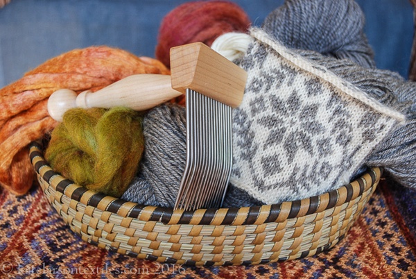 Basket with wool comb, spinning fibers, gray yarn, and a white and gray stranded knitting swatch in a star pattern. The basket is on a multicolor diamond twill mat.